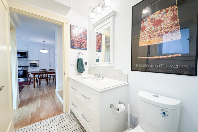 White bathroom vanity with marble countertop and framed art, opening to a dining area with hardwood floors.
