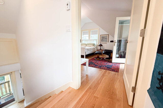 Hallway with light hardwood floors opening into a loft bedroom with a red patterned rug and slanted ceiling.