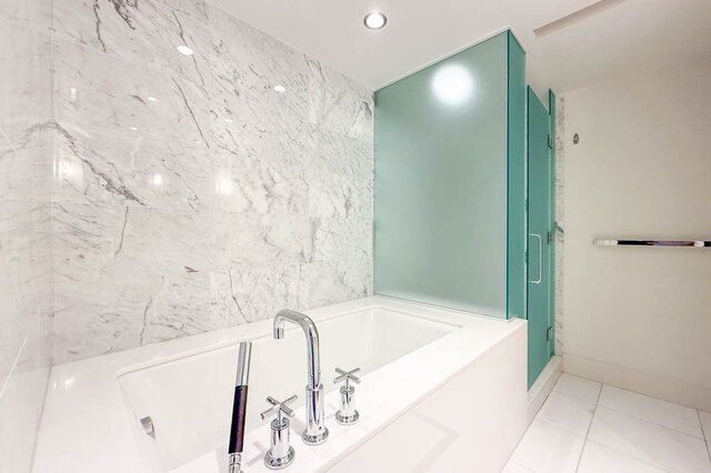 Modern bathroom with white marble tile walls, a built-in tub with chrome fixtures, and a frosted green glass shower.