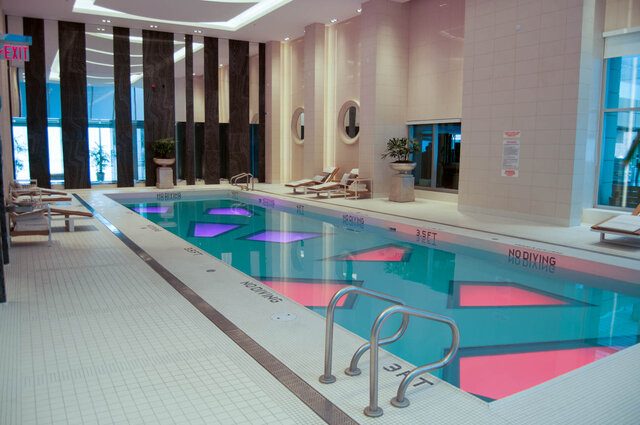 Spacious indoor pool area with turquoise water, white tiled deck, lounge chairs, and dark wood columns. - 2702-667 Howe Street, Vancouver Downtown