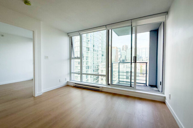 Furnished living room with gray sectional sofa and hardwood floors, opening to a white kitchen and dining nook.
