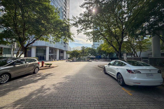 Paved courtyard flanked by mature trees and a modern glass condo building, with two parked cars. - 1603-668 Citadel Parade, Vancouver Downtown