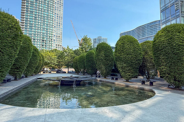 Outdoor courtyard with a central reflecting pool and rows of manicured topiary trees framed by high-rise towers.