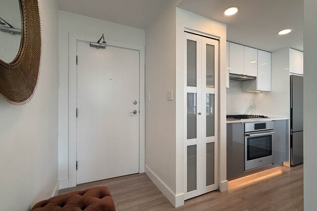 Entryway with light wood flooring and frosted glass closet doors opening to a white kitchen with stainless steel oven.