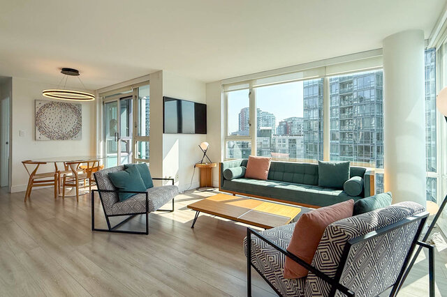 Bright living room with hardwood floors, green sofa, and large windows framing a downtown city view.