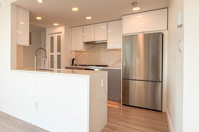 Modern kitchen with white glossy cabinets, marble island, and stainless steel refrigerator on light hardwood floors.