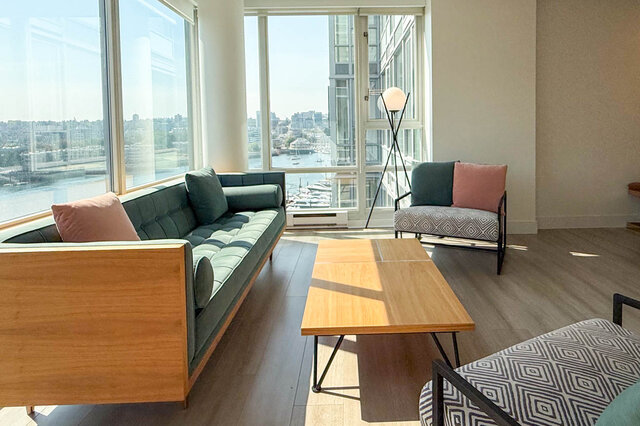 Bright living room with hardwood floors, a green sofa, and floor-to-ceiling windows framing a marina city view.