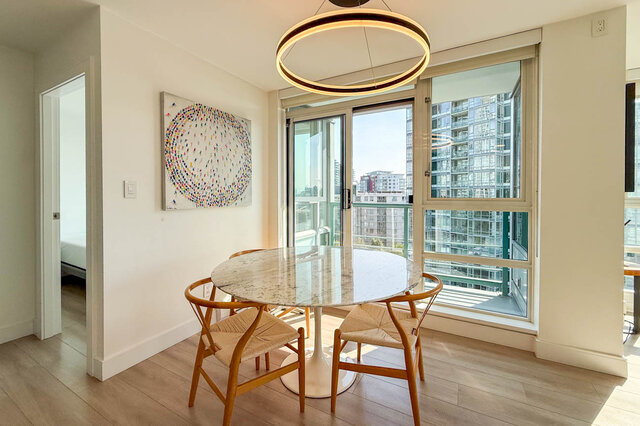 Bright dining nook with a round marble table, woven wood chairs, and floor-to-ceiling windows framing a city view.