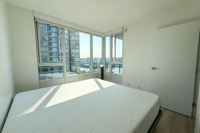 Bright bedroom with corner windows offering a city and water view, light wood floors, and a white bed.