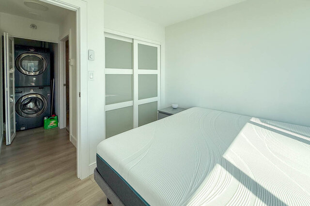 Bedroom with a made bed, light wood-look floors, and an in-unit stacked washer/dryer visible through an open doorway.