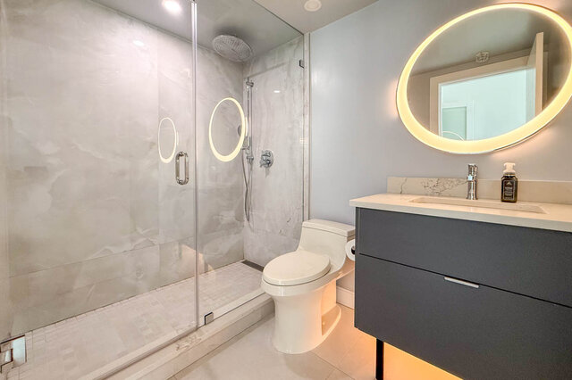 Modern bathroom featuring a glass-enclosed walk-in shower with marble tile and a floating vanity under a round backlit...