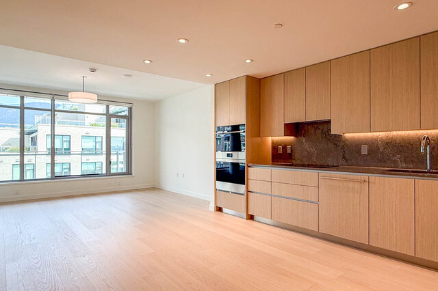 Kitchen with light wood cabinetry, dark stone backsplash, and hardwood floors beside a large window. - 607-777 Harbour Way, North Vancouver
