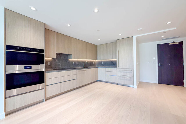 Modern kitchen with light wood cabinetry, dark stone countertops, and pale hardwood floors. - 607-777 Harbour Way, North Vancouver
