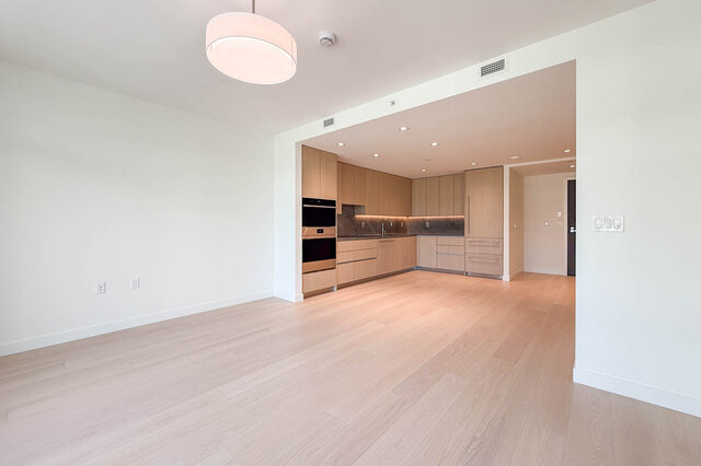 Open living area with light hardwood floors and an adjacent kitchen featuring wood cabinetry and dark stone countertops.