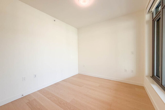 Empty bedroom with light hardwood floors and crisp white walls, featuring a window and recessed ceiling light. - 607-777 Harbour Way, North Vancouver