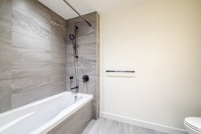Modern bathroom with grey marble-look tile walls, a white bathtub, and dark chrome shower fixtures. - 607-777 Harbour Way, North Vancouver