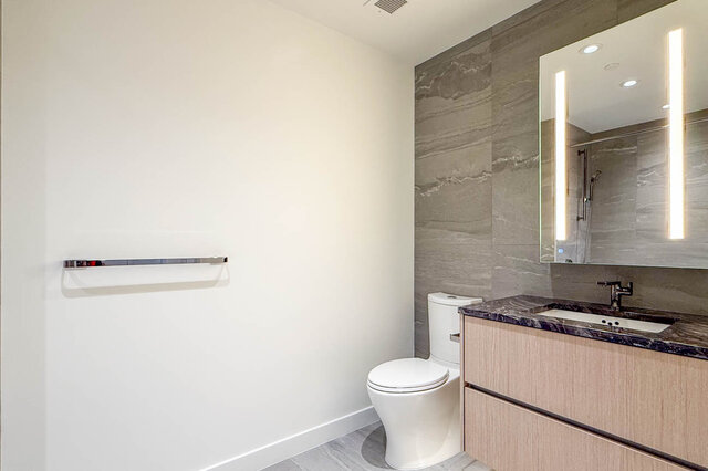 Modern bathroom with light wood vanity, dark granite countertop, and a backlit mirror against gray marble tile. - 607-777 Harbour Way, North Vancouver