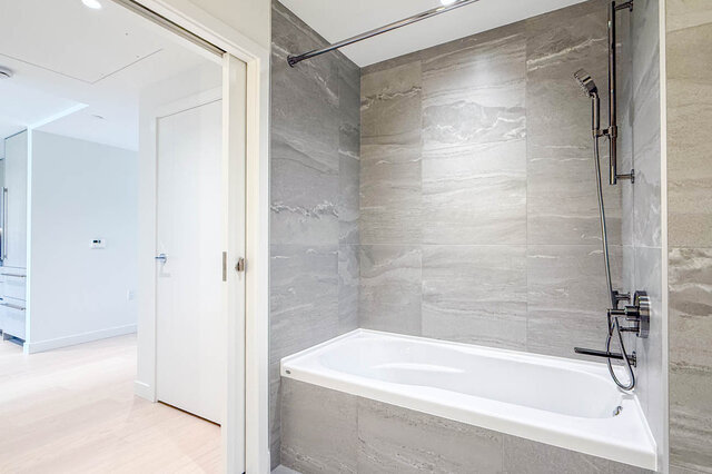 Bathroom featuring a white soaking tub set into gray marble-look tile walls with chrome shower fixtures. - 607-777 Harbour Way, North Vancouver
