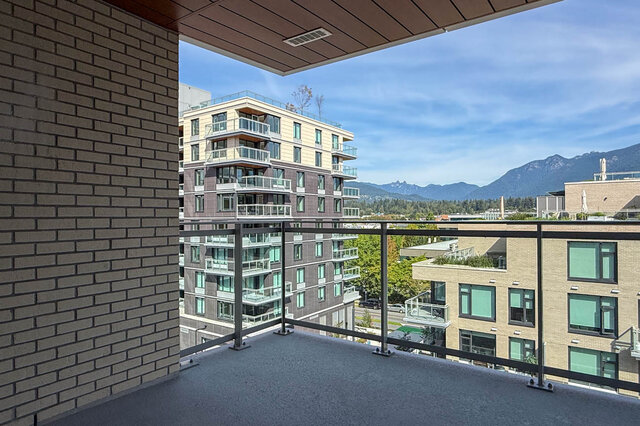 Private balcony with a brick accent wall and glass railing framing a distant mountain view. - 607-777 Harbour Way, North Vancouver