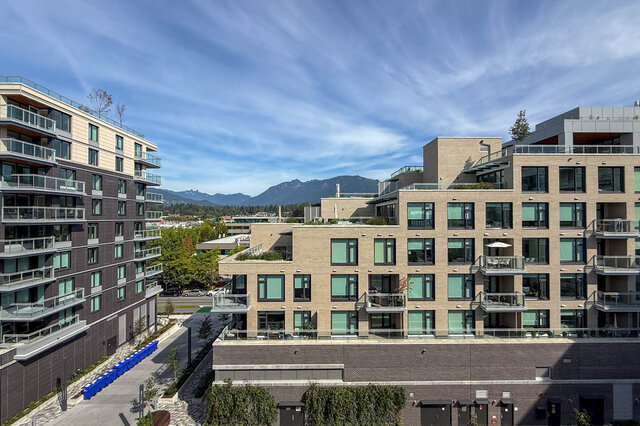 Exterior view of modern condo with glass-railed balconies and a distant mountain backdrop. - 607-777 Harbour Way, North Vancouver