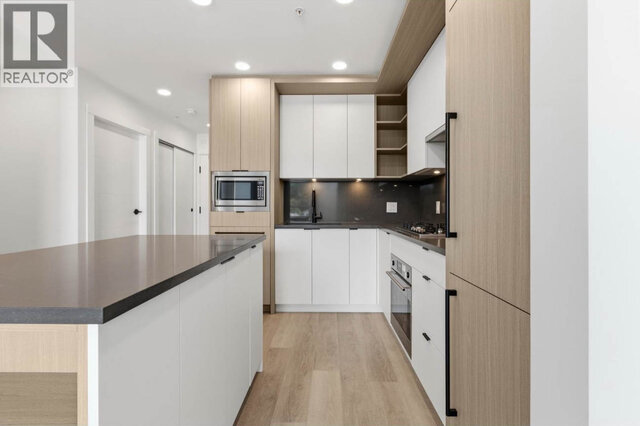 The kitchen, a dark-topped island in front of white flat-panel cabinets and light wood-tone tall cabinets, a built-in microwa