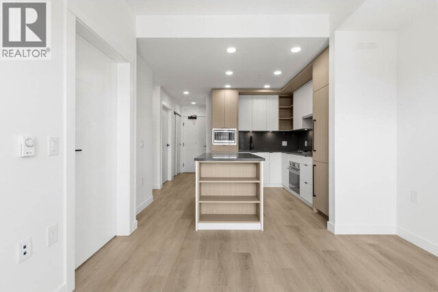 The kitchen island with open shelving on its end, the hall to the entry door and closet doors beyond, recessed lighting acros