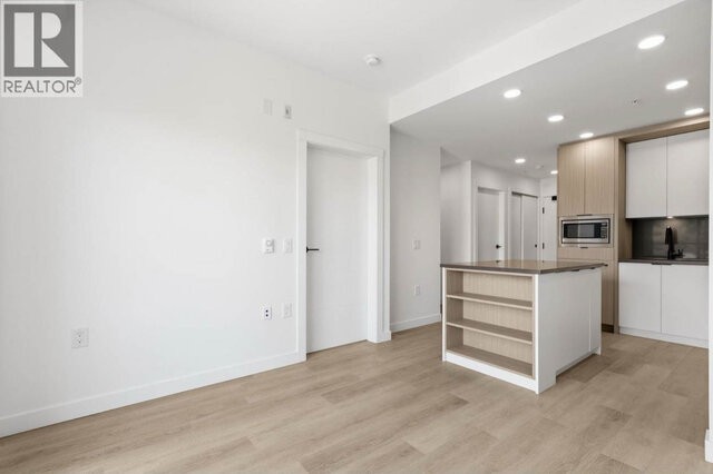 The empty open-plan living area facing the kitchen island, white walls and light vinyl plank floors, the bedroom door to the 
