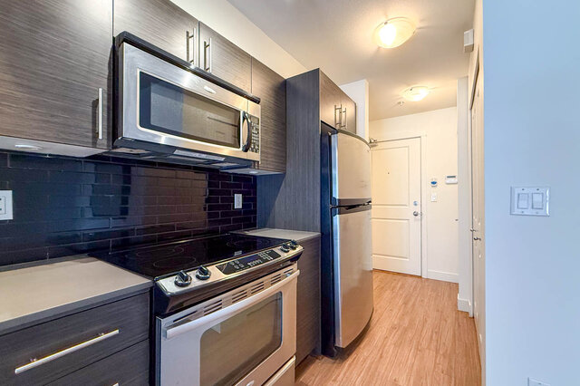 Kitchen with dark wood-grain cabinets, stainless over-range microwave, range and fridge, glossy black subway tile, and the fr