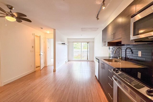 Long open living space with hardwood floors and a ceiling fan, the kitchen along one wall and a sliding door to the balcony a