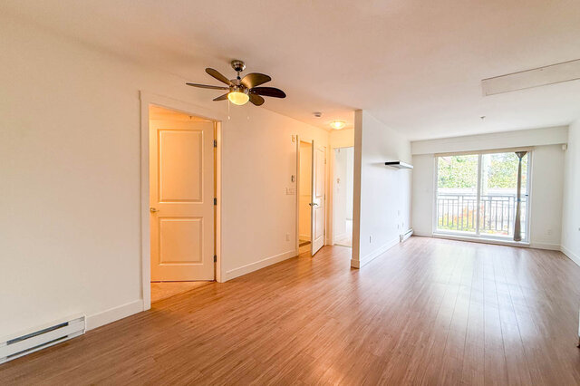 Bright empty living room with hardwood floors, a ceiling fan, doors to the den and bathroom, and a sliding door to the balcon