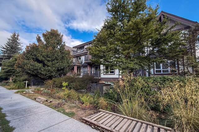 Three-storey craftsman-style building with balconies behind gardens of tall grasses and trees along the sidewalk.