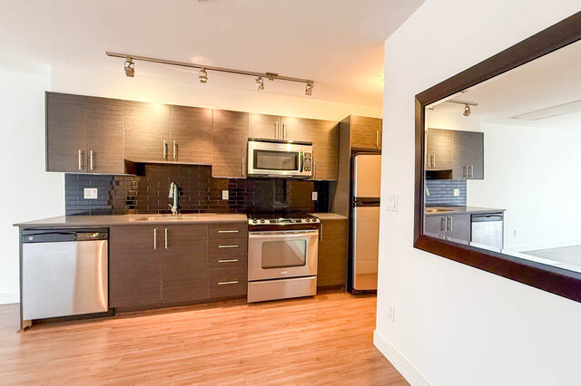 Full kitchen wall with dark wood-grain cabinets, stainless dishwasher, range, microwave and fridge, glossy black subway tile 
