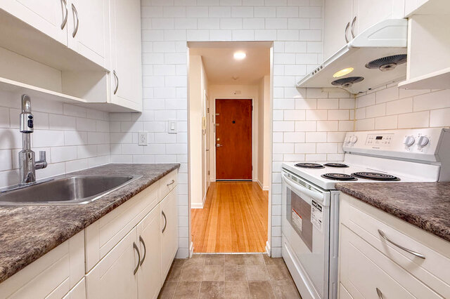 The galley kitchen with a white stove under a range hood and subway tile walls, looking out to the front door