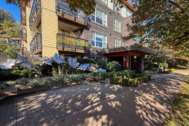 Building front with metal leaf sculptures in the garden beds and a covered entry, under trees. - 402-3205 Mountain Highway, North Vancouver
