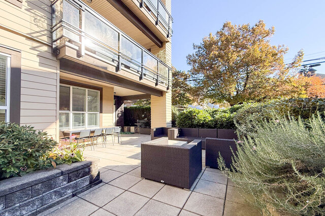 Shared courtyard with wicker seating, a long outdoor table and planters under the trees. - 402-3205 Mountain Highway, North Vancouver