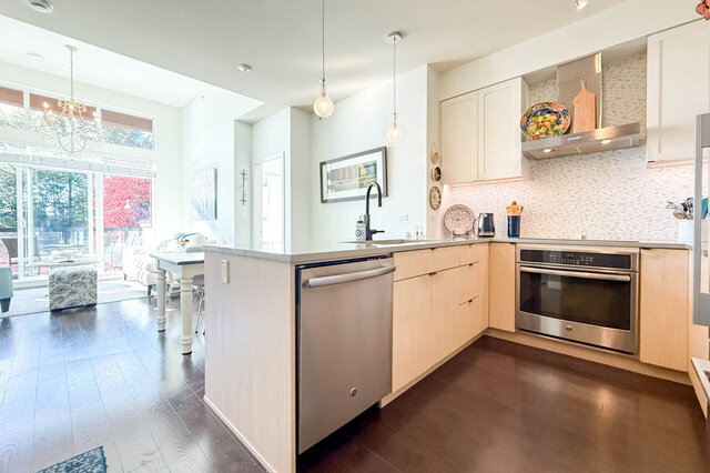 Kitchen with stainless appliances, a wall oven, a tile backsplash and pendant lights, open to the living room and its tall wi