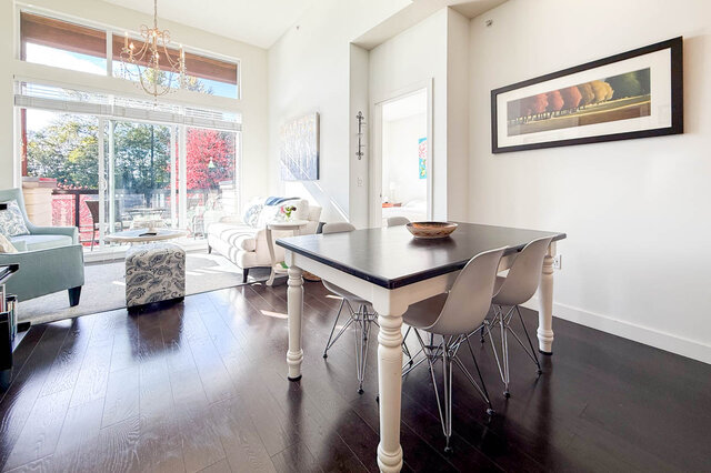 Dining table for four next to the living room, a chandelier and tall windows beyond, a bedroom door open behind.