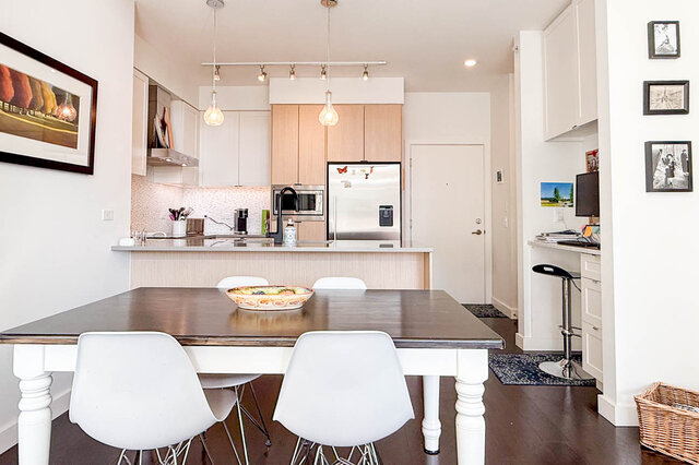 Dining table in front of the kitchen peninsula, stainless fridge and microwave, the front door beside it. - 402-3205 Mountain Highway, North Vancouver