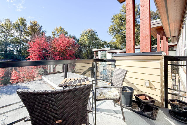 Private balcony with wicker chairs and a bistro table, glass railing and red autumn trees beyond. - 402-3205 Mountain Highway, North Vancouver