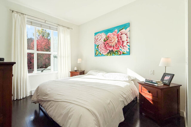 Primary bedroom with a made-up queen bed, two nightstands, a dresser and a big window with red autumn trees outside.