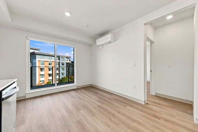 Empty living area with the sliding glass door to the balcony and a wall air conditioner, the hall to a bedroom on the right.