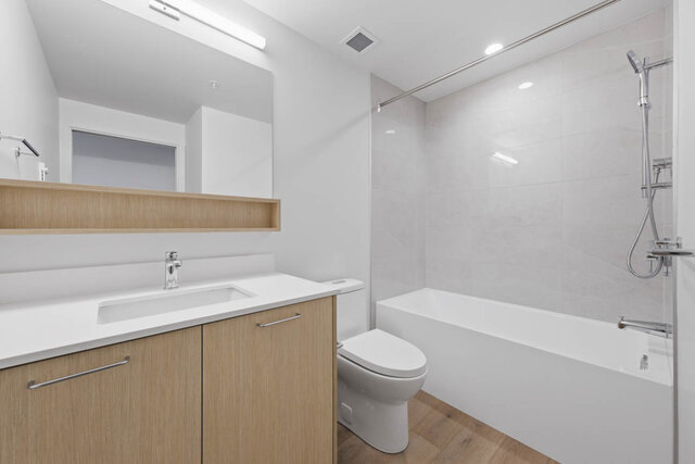 Main bathroom with a tub and shower, a tile surround, a light wood vanity with a quartz counter and an open shelf under the m