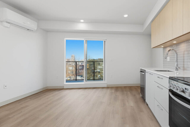 Living area with the sliding glass door to the balcony, a wall air conditioner and the kitchen sink and range on the right.