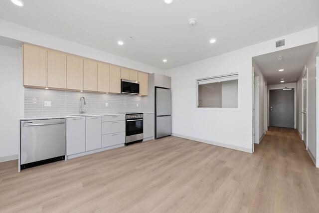 Kitchen along the wall with stainless appliances, an interior window beside it and the hall leading back to the entry door.
