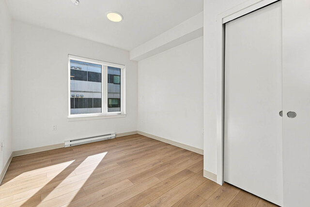Empty primary bedroom with a window, a sliding closet and sun across the hardwood floors. - 405-1529 Oxford Street, North Vancouver