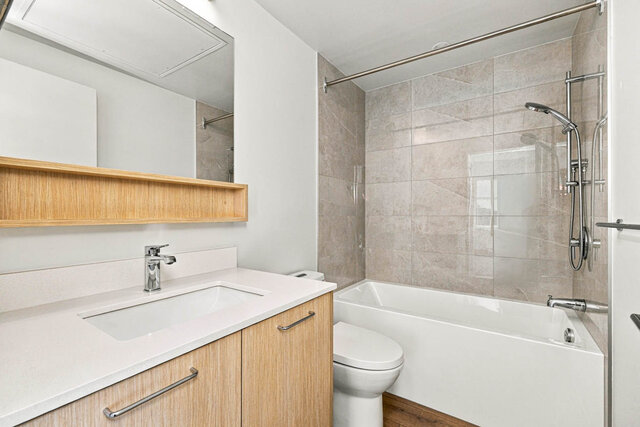 Full bathroom with a tub and shower, a tile surround and a light wood vanity with a quartz counter under a mirror and open sh