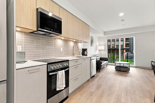 Galley kitchen with light oak upper cabinets, a quartz counter, a tile backsplash, under-cabinet lighting, a stainless range,