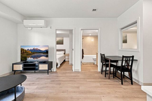 Living room with the TV on its media console, the dining table for two, and doors to the second bedroom and a bathroom side b