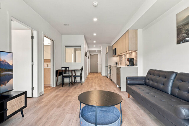 Living room with the leather sofa and round coffee table, looking past the dining table and kitchen to the entry door.