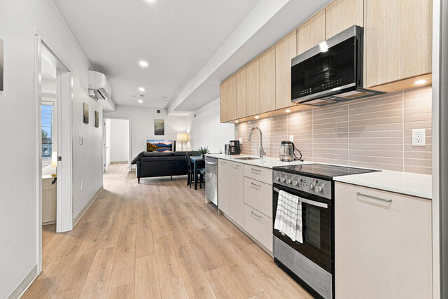 Galley kitchen running down to the furnished living area with a leather sofa, TV and dining table, past an open bedroom door 